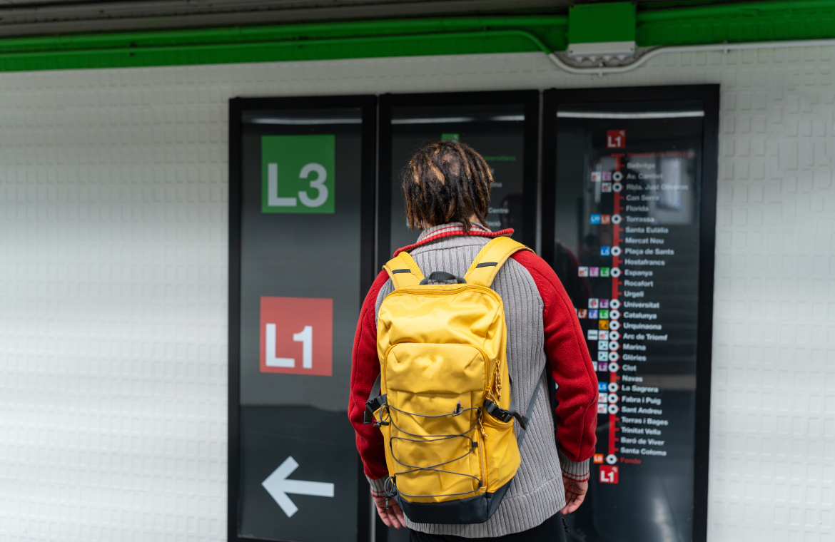 A confused young Black man with braids and a yellow backpack checking the red and green line metro signs at Plaça Espanya.