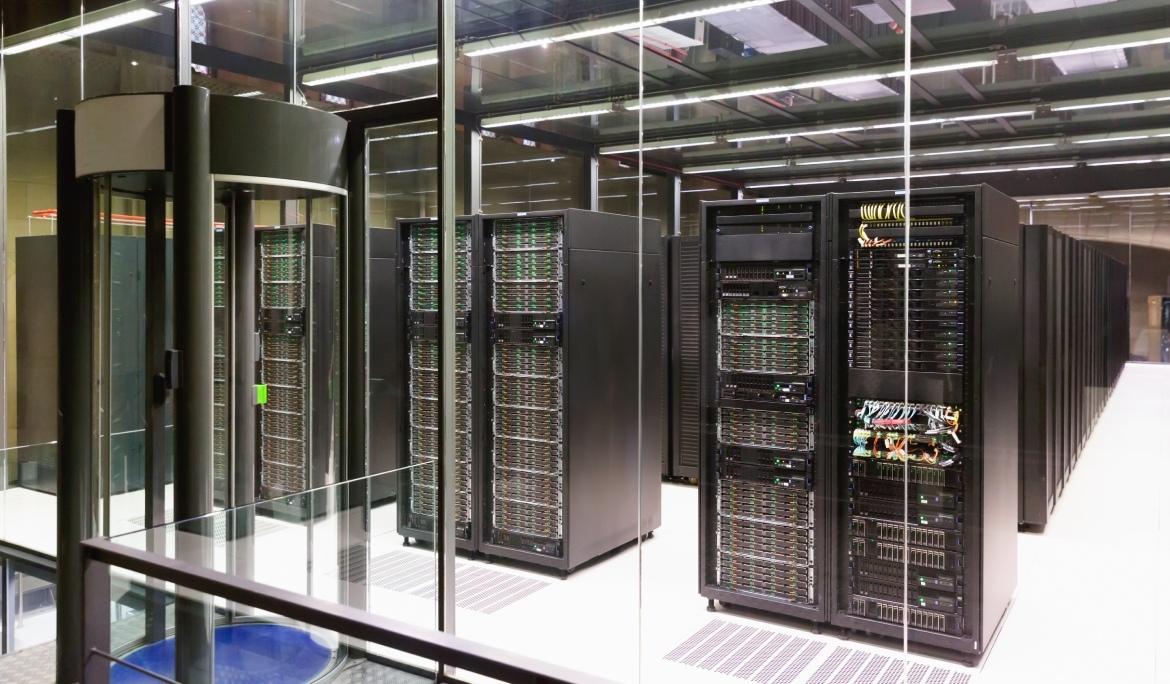Image of the Supercomputing Center showing rows of supercomputers with cables, buttons and indicator lights behind a glass panel and a security door. It represents one of Barcelona’s key technological hubs.