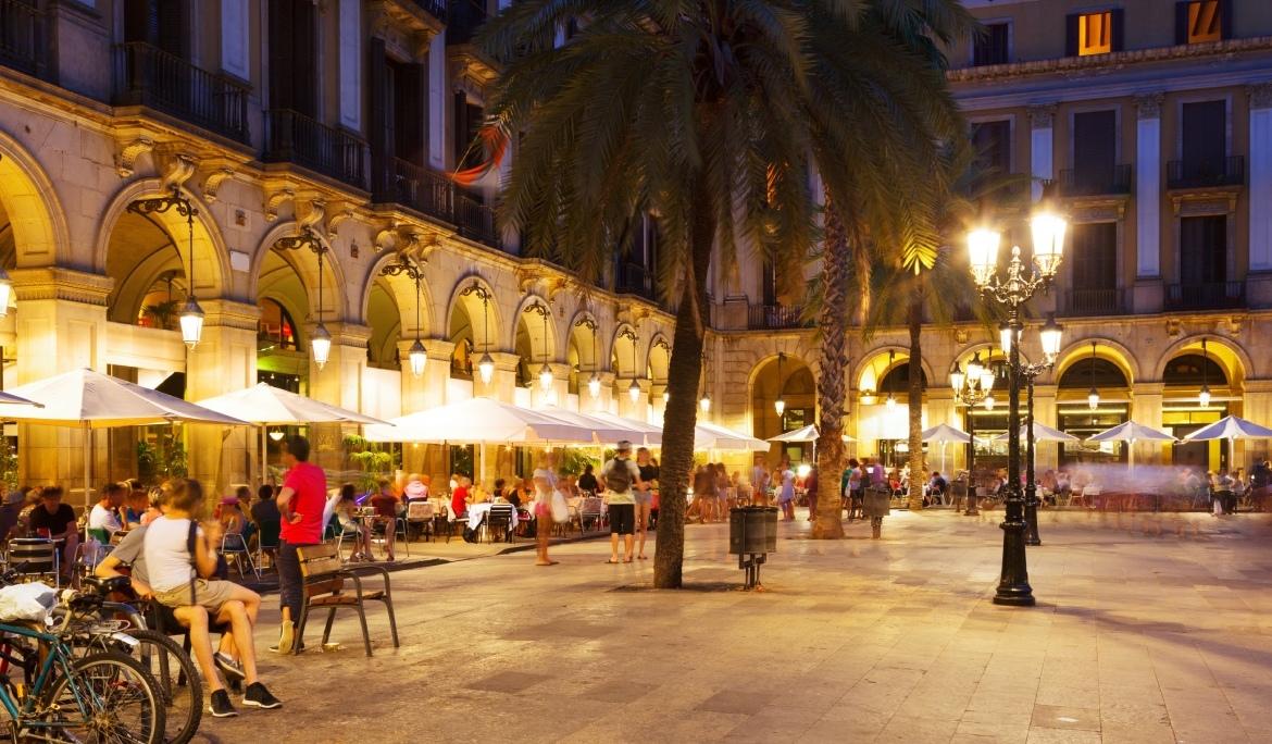 A nighttime view of Barcelona with a lively square full of bars and outdoor tables, where people enjoy dinner in the heart of the city.