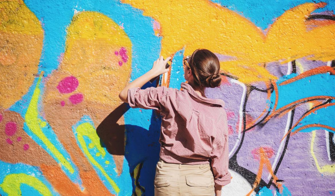 Image of a female street‑art artist creating a graffiti piece. She’s dressed casually, with a slightly messy bun, spray‑painting a colourful wall.