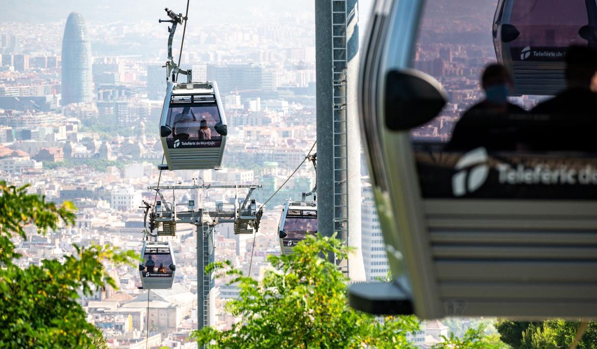 Image of a red cable car cabin hovering over the green landscape of Montjuïc hill with the city in the background.