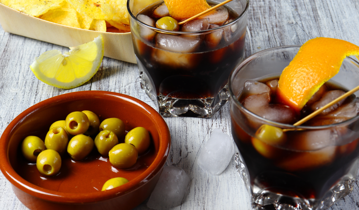 Una taula amb un platet d'olives i dos gots amb vermut i un tall de taronja.