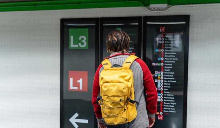 A confused young Black man with braids and a yellow backpack checking the red and green line metro signs at Plaça Espanya.